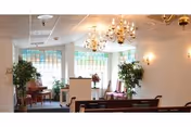 A bright and elegant common area with large windows, chandeliers, wall sconces, and several potted plants. There are wooden benches and chairs arranged facing a podium, suggesting a space for gatherings or presentations.