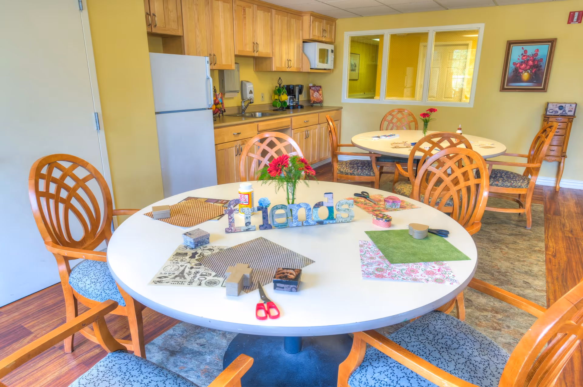 A bright room with two round tables surrounded by wooden chairs with patterned cushions. The table in the foreground has craft supplies including scissors, patterned paper, glue, and decorative letters spelling 'friends'. The background shows a kitchenette with wooden cabinets, a refrigerator, a microwave, and a coffee maker. The walls are painted yellow and there is a framed floral painting on the wall.