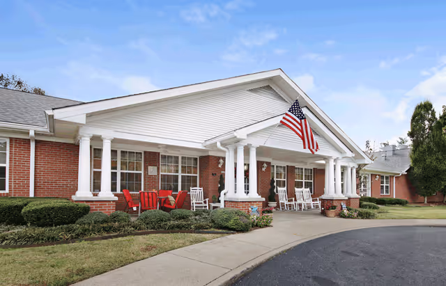 Front exterior view of a single-story brick building with white columns and a covered porch. The porch has white rocking chairs and red Adirondack chairs. An American flag is mounted on the front of the building. There are shrubs and a curved driveway in front of the building under a partly cloudy sky.