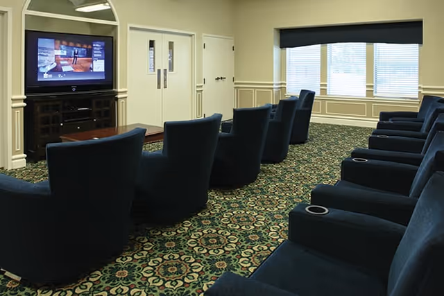 Rows of blue upholstered armchairs in a carpeted TV lounge facing a large flat-screen television and windows.