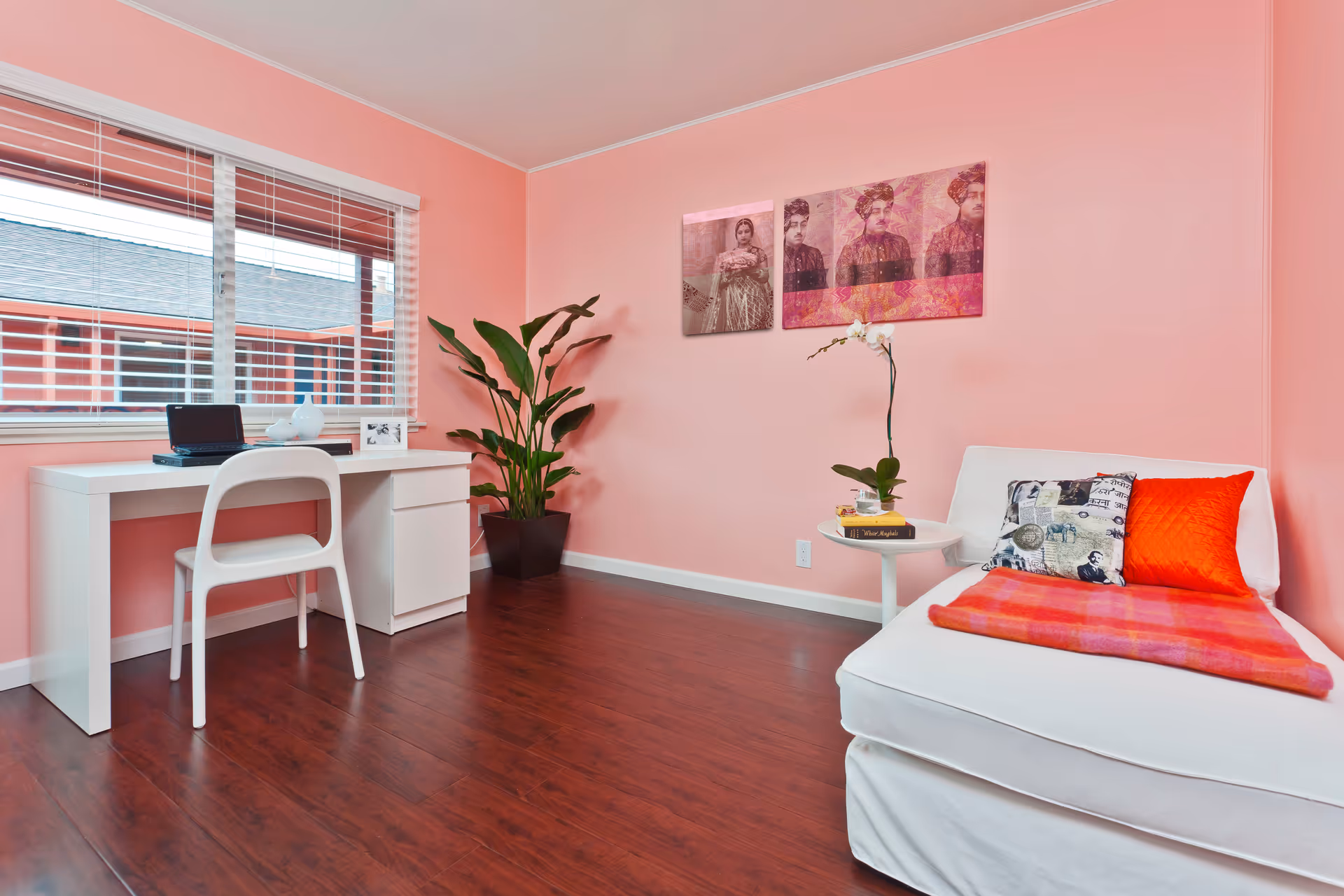 A cozy room with pink walls and wooden flooring featuring a white desk and chair near a window with blinds. There is a large green potted plant in the corner, and a white cushioned chair with colorful pillows and a red blanket. A small round table next to the chair holds a potted orchid and some books. Two framed artworks hang on the wall above the chair.