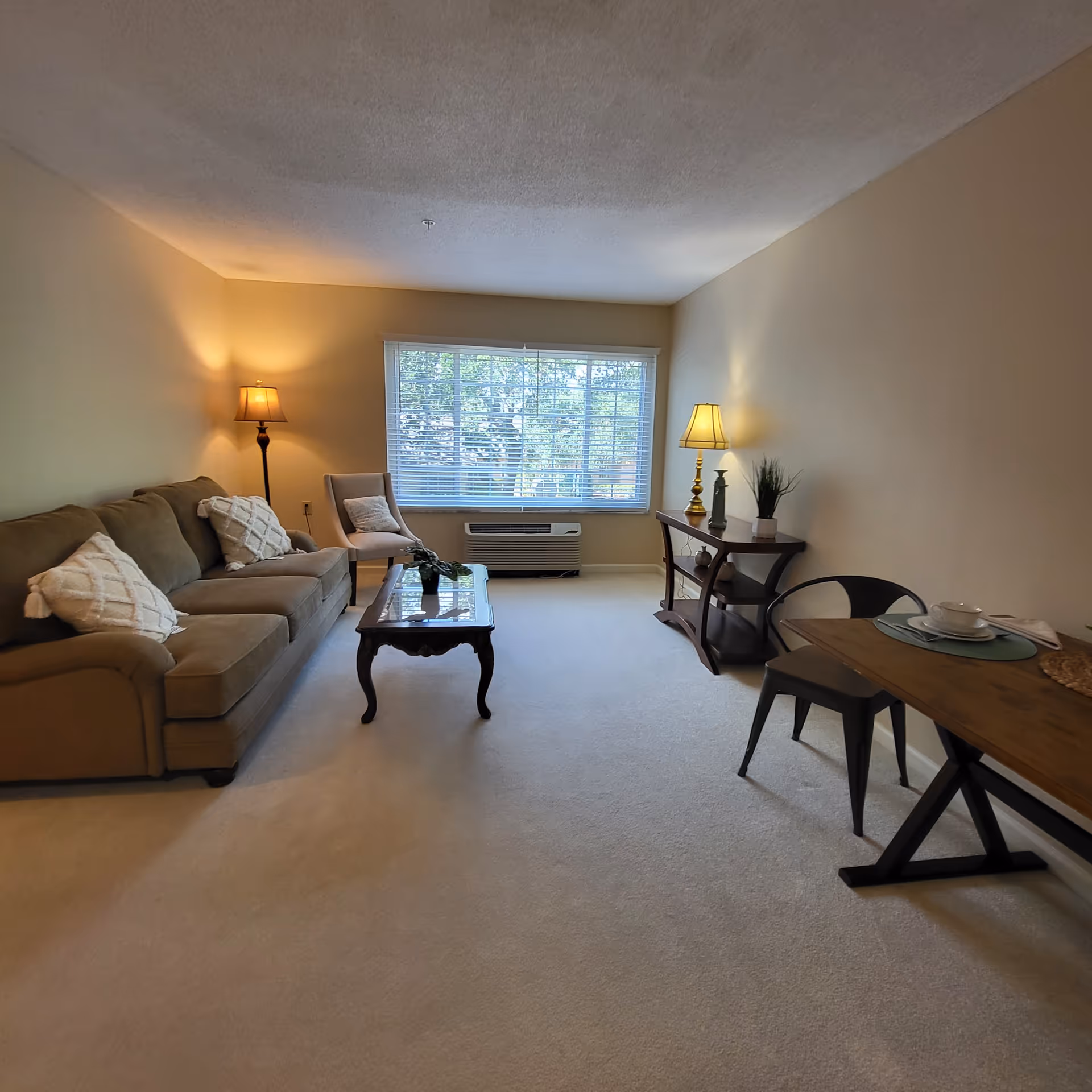 A cozy living room with beige walls and carpet. The room features a brown sofa with two white textured pillows, a wooden coffee table with a small plant, a beige armchair, and a wooden console table with a lamp, decorative items, and a small plant. A wooden dining table with a place setting and two metal chairs is positioned on the right side. A large window with white blinds lets in natural light, and there is an air conditioning unit below the window.
