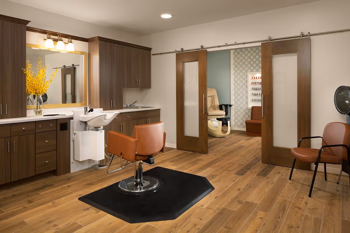Interior view of a salon area in a senior living facility featuring a brown salon chair in front of a white sink and wooden cabinetry with a large mirror above. The room has wooden flooring and sliding wooden doors leading to another room with a beige pedicure chair and a nail polish display on the wall. There is also a brown chair with armrests and a hair dryer on the right side.