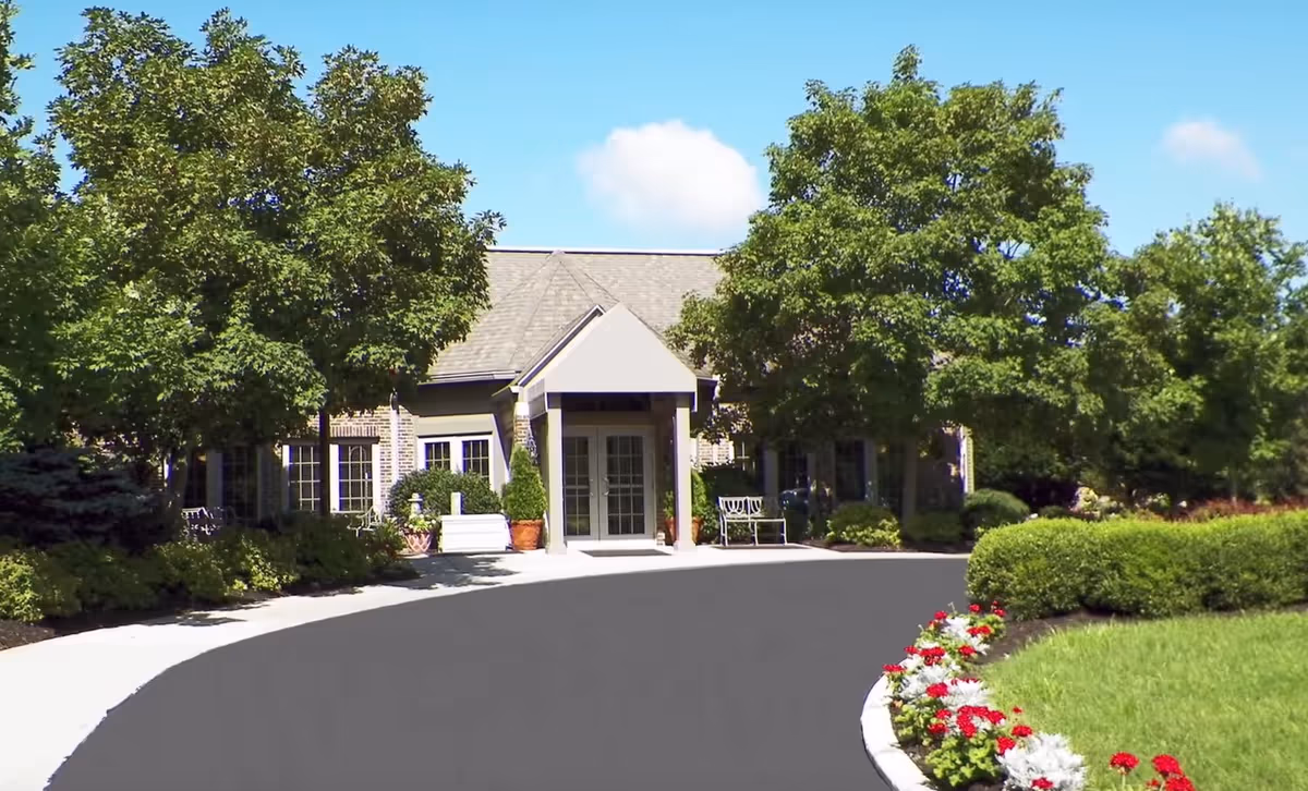 Front exterior view of Burton's Ridge Assisted Living facility with a paved driveway, green trees, bushes, and a flower bed with red and white flowers under a clear blue sky.