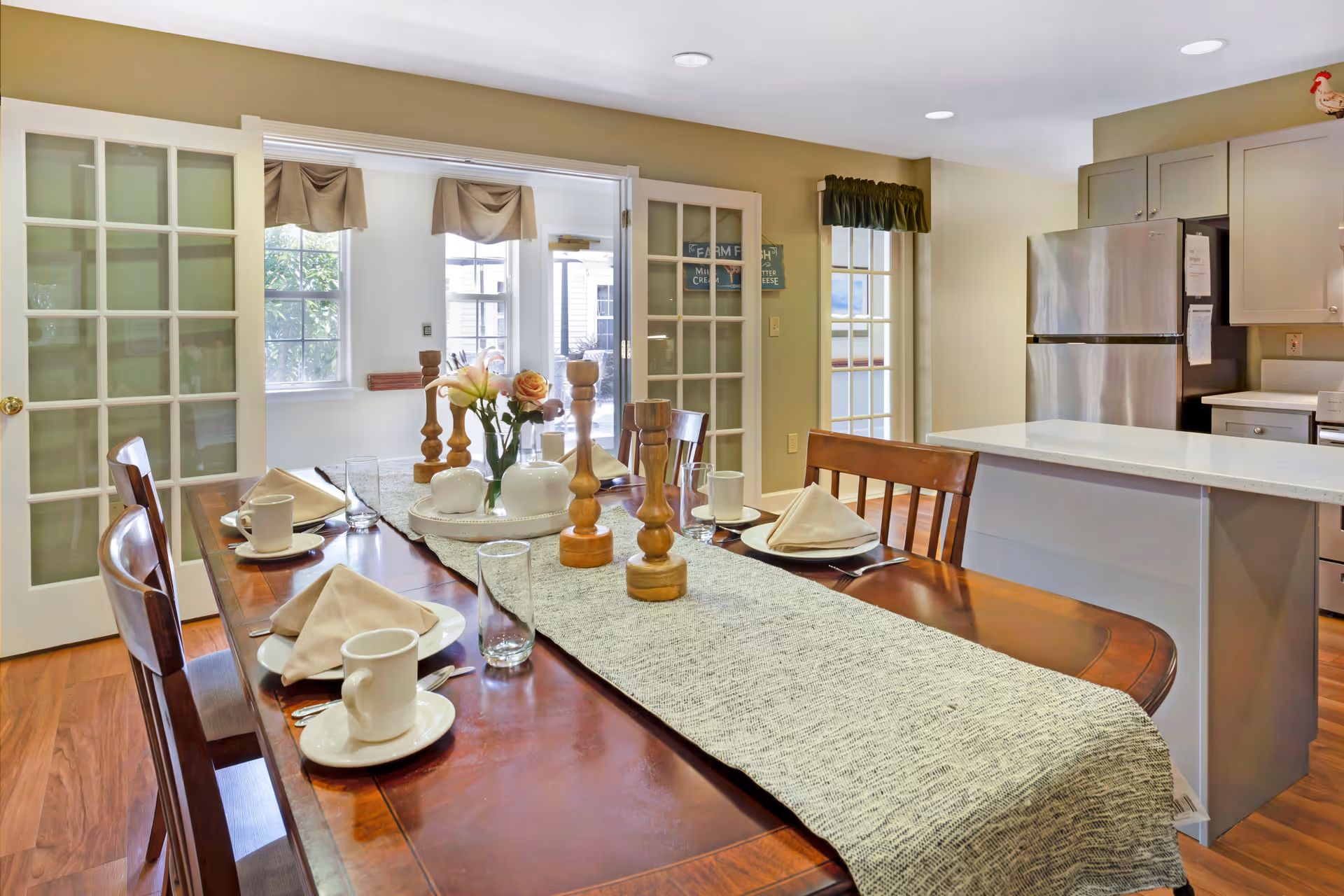 A dining area with a wooden table set for four, featuring white plates, cups, napkins, and glassware. The table has a gray runner and decorative wooden candle holders and a vase with flowers. In the background, there is a kitchen with stainless steel refrigerator, light-colored cabinets, and a countertop. French doors and windows allow natural light into the space.