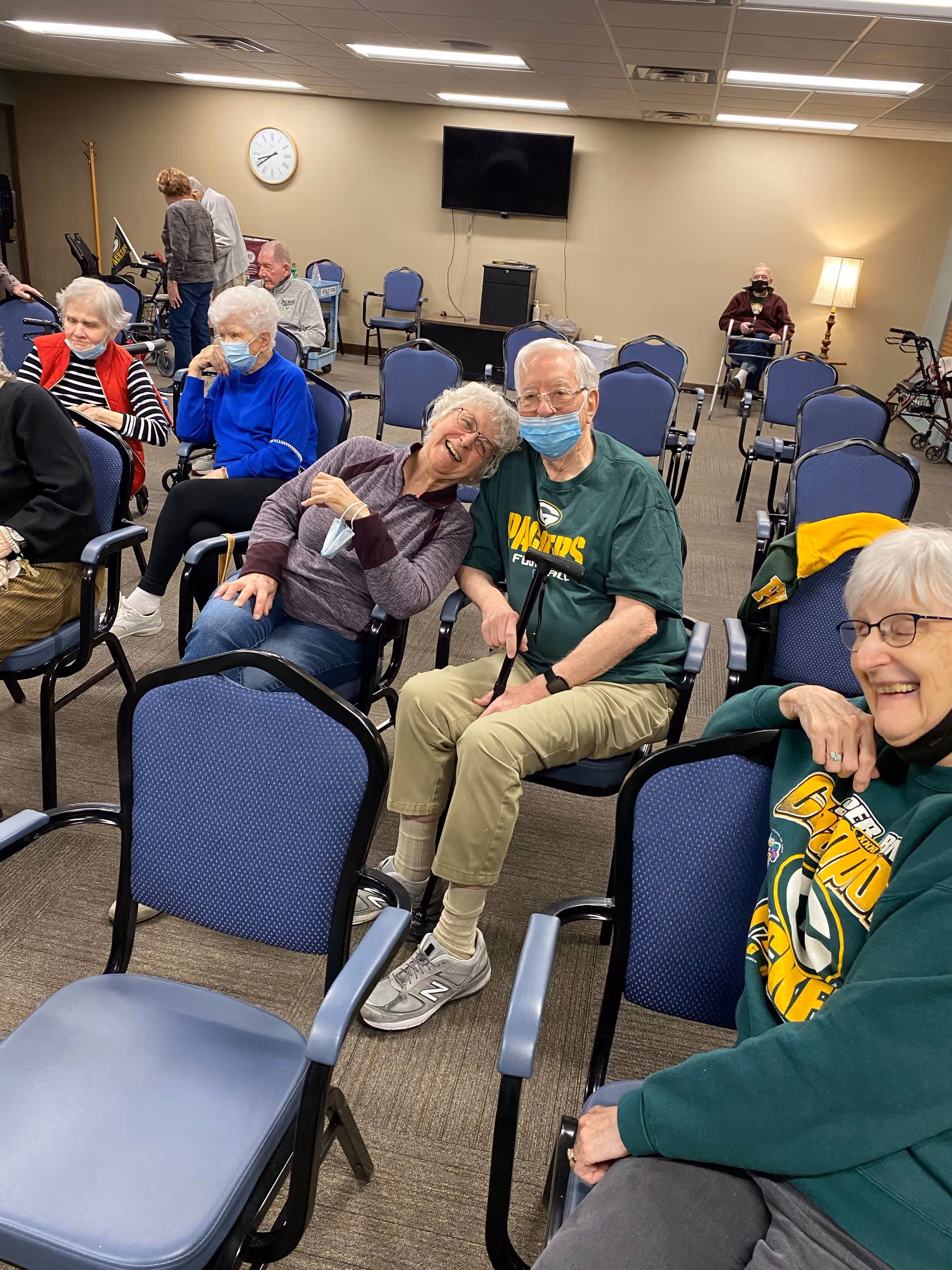 A group of elderly people sitting in a community room with blue chairs. Some are wearing masks, and a few are smiling and interacting. The room has beige walls, a clock, a flat-screen TV mounted on the wall, and a standing lamp in the corner.