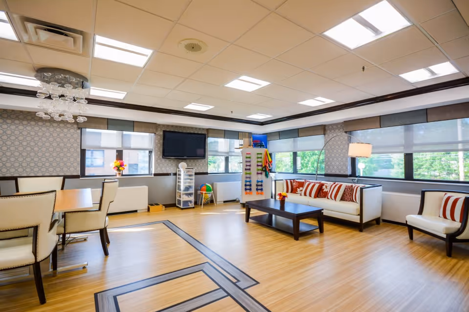 A bright and spacious common area with large windows letting in natural light. The room features a white sofa with red and white striped cushions, a matching armchair, a dark wooden coffee table, and a dining table with cream-colored chairs. There is a flat-screen TV mounted on the wall, a floor lamp, and a storage unit with colorful exercise bands and a beach ball. The floor has a light wood finish with a decorative inlay, and the ceiling has recessed lighting and a modern chandelier.