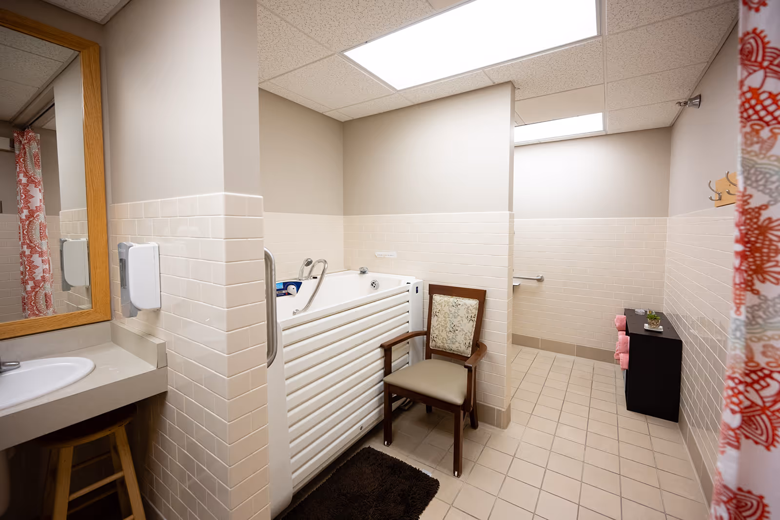Accessible tiled bathroom featuring a walk-in bathtub with grab bars, a chair, sink with mirror, and a small storage cabinet.