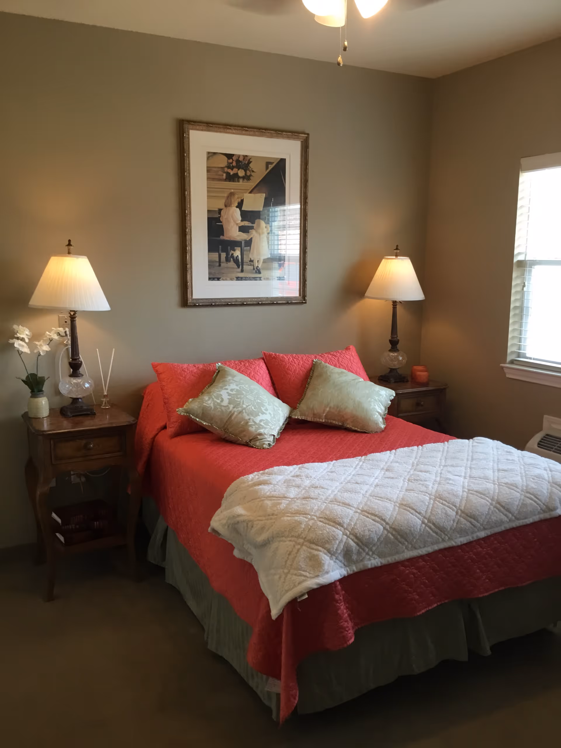 Neatly made bed with a red bedspread, green pillows, bedside tables and lamps in a warmly lit bedroom.