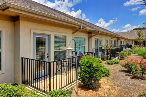 Exterior view of a senior living facility showing a row of ground-level apartment units with small fenced patios, greenery, and flowering bushes under a partly cloudy sky.