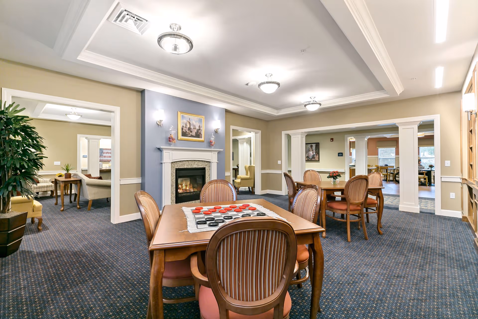 A well-lit common area in a senior living facility featuring a wooden table with chairs arranged around it. A checkers game is set up on the table. The room has a fireplace with a painting above it, beige walls with white trim, and blue carpeting. Adjacent rooms with additional seating and tables are visible through open doorways.