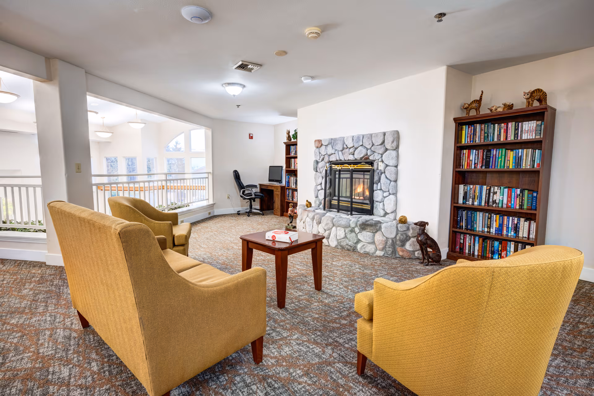 A cozy common area with a stone fireplace, two yellow armchairs, a yellow sofa, a wooden coffee table with a book on it, a bookshelf filled with books, and a desk with a computer and office chair near a window.