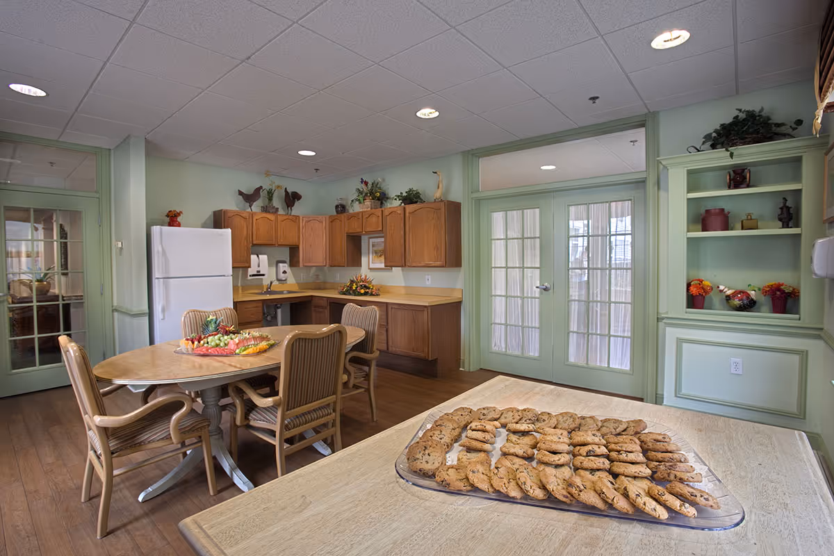 A cozy dining area with a wooden table surrounded by four chairs, a platter of cookies on a nearby counter, a kitchen area with wooden cabinets, a white refrigerator, and decorative plants and items on shelves and countertops. The room has light green walls and double glass doors with curtains.