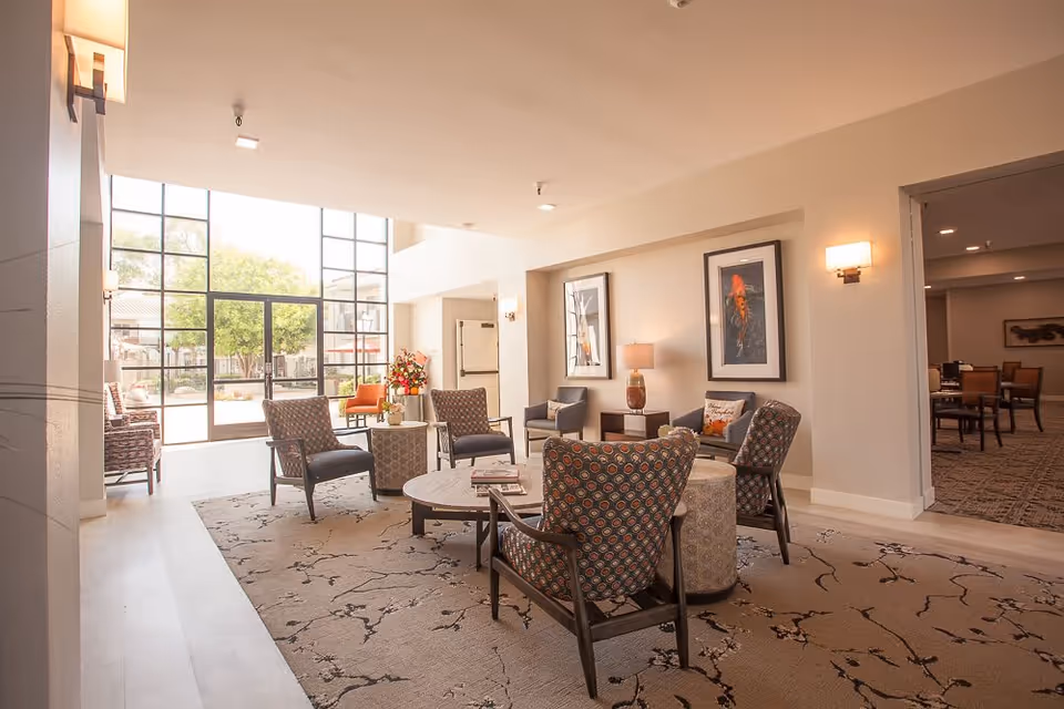 A bright and spacious senior living facility common area with large floor-to-ceiling windows letting in natural light. The room features several patterned armchairs arranged around a circular coffee table on a decorative rug. There are framed artworks on the walls, a table lamp, and a flower arrangement near the entrance to an outdoor patio area. In the background, there is a dining area with tables and chairs.