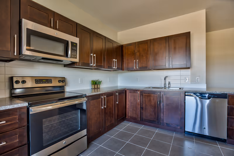 Modern kitchen with dark wood cabinets, stainless steel appliances including a microwave, oven, and dishwasher, a double sink, and gray tiled floor.