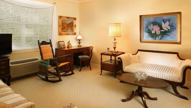 A cozy living room with beige walls and carpet, featuring a wooden rocking chair with a green and red blanket draped over it, a wooden desk with a small lamp and framed pictures, a side table with a lamp, a cream-colored upholstered sofa, and a wooden coffee table with a glass ashtray. Two framed paintings hang on the walls, and a window with white blinds lets in natural light.