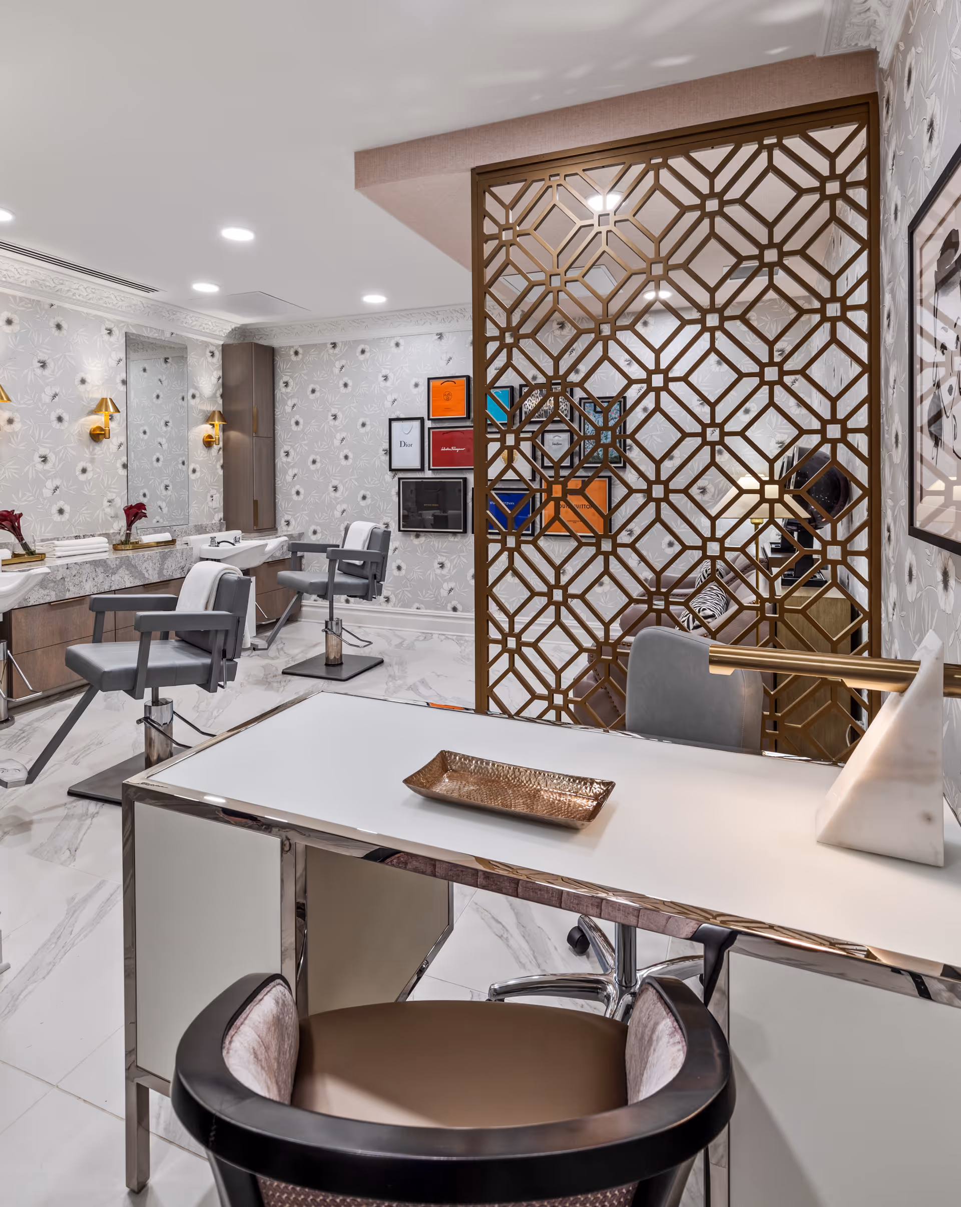 Interior of a stylish salon area with two gray salon chairs facing mirrors on a floral patterned wall. A decorative gold geometric room divider separates the salon area from a white desk with a modern lamp and a bronze tray. The floor is white marble with gray veining, and framed artwork is visible on the walls.