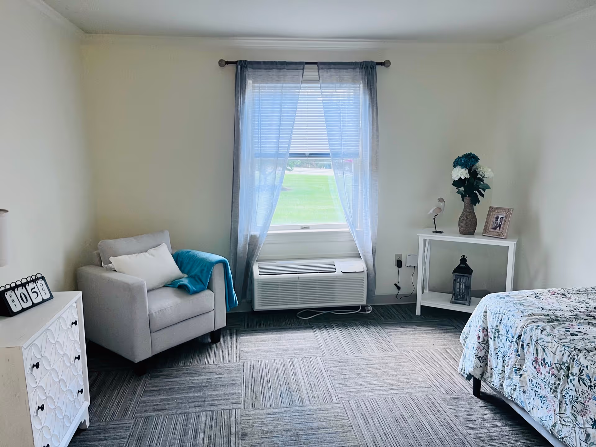 A bright and cozy bedroom with a window covered by sheer curtains, a light gray armchair with a white pillow and blue throw blanket, a white cabinet with a calendar showing October 5th, and a white side table holding a vase with flowers, a small bird figurine, a framed photo, and a lantern. The bed has a floral patterned bedspread.
