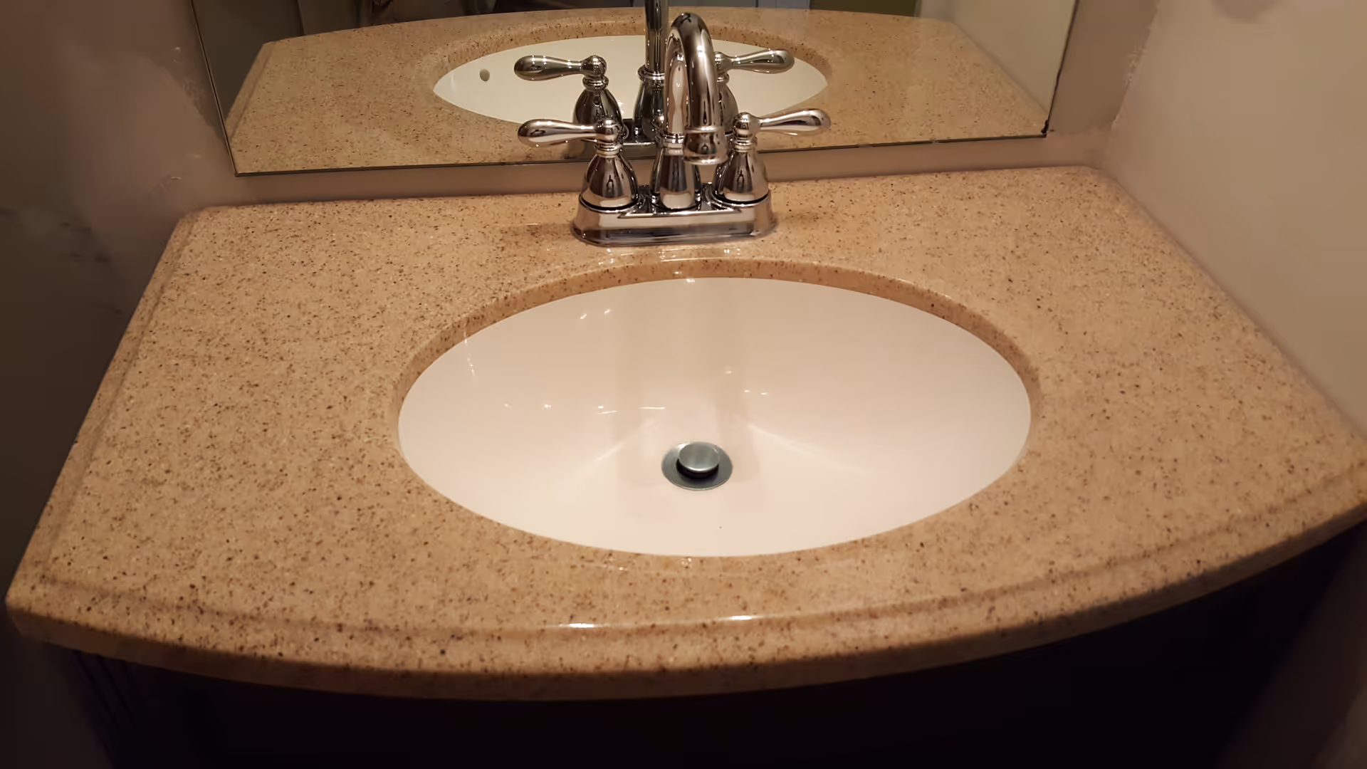 A bathroom sink with a beige speckled countertop and a chrome faucet. A mirror is mounted on the wall behind the sink.