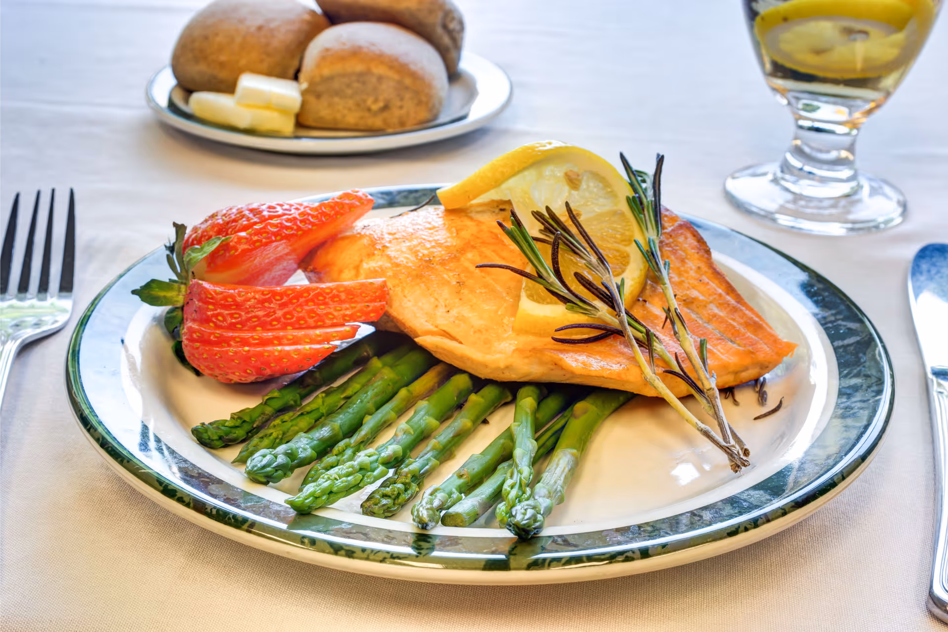 A plated meal featuring a piece of cooked salmon garnished with lemon slices and rosemary, served on a bed of asparagus with sliced strawberries on the side. In the background, there is a plate with bread rolls and butter, and a glass of a light-colored beverage on a white tablecloth.