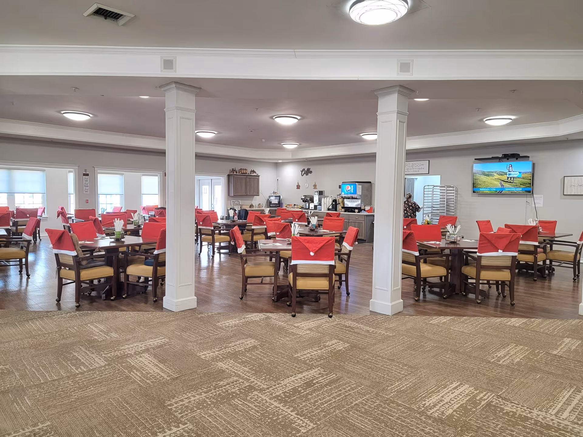 Large dining room with multiple round tables and chairs draped in red napkins, central columns, and a TV on the wall.