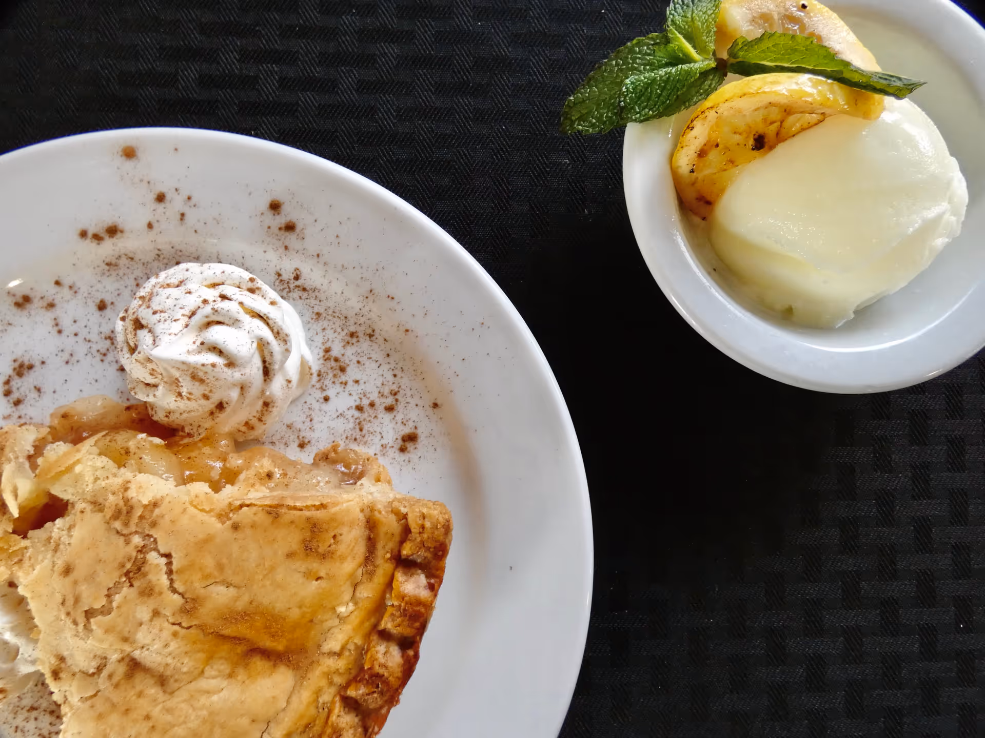 A close-up view of a slice of apple pie with a dollop of whipped cream sprinkled with cinnamon on a white plate, accompanied by a small bowl of vanilla ice cream garnished with a mint leaf and a slice of caramelized banana, all placed on a black textured surface.