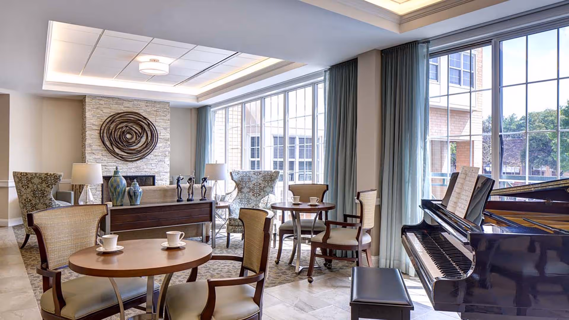 A bright and elegant common area with large windows letting in natural light, featuring a grand piano with sheet music, several round tables with chairs, two armchairs near a stone fireplace adorned with decorative vases and sculptures, and light blue curtains.