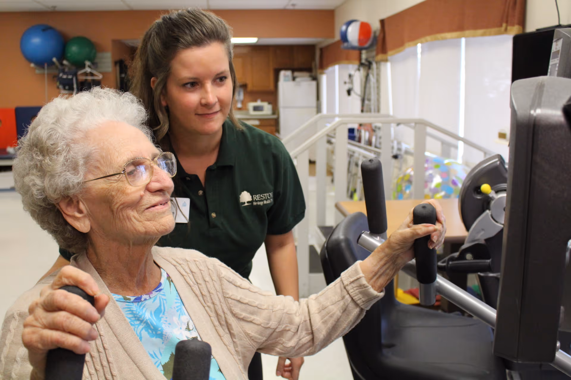 An elderly woman using exercise equipment in a rehabilitation or therapy room, assisted by a younger female staff member wearing a green shirt with a name tag. The room contains various therapy and exercise equipment in the background.