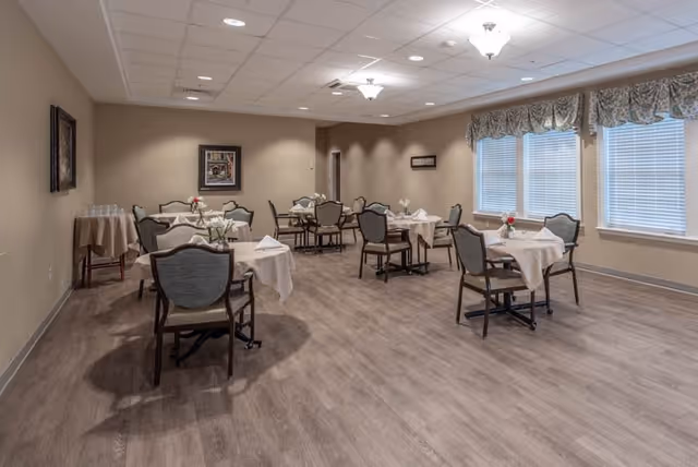 A dining room in an assisted living facility with several tables covered in white tablecloths, each set with napkins and small flower arrangements. The room has wood flooring, beige walls, framed artwork, and large windows with patterned valances allowing natural light to enter.