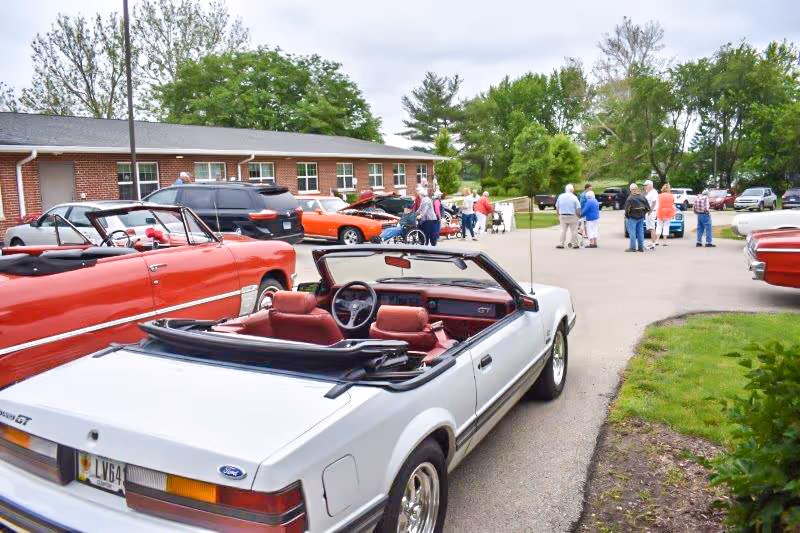 A group of people and classic cars gathered in a parking lot outside a low brick care facility building.