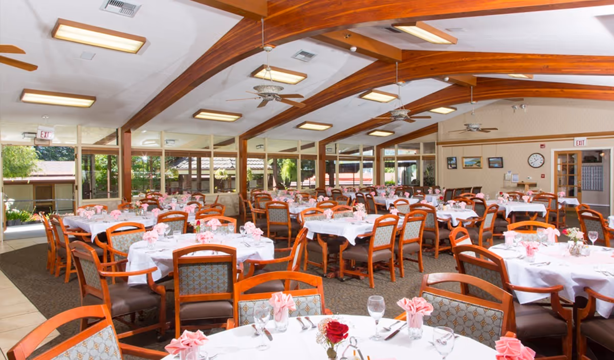 A spacious dining room with multiple round tables covered in white tablecloths, each set with pink folded napkins, glassware, and silverware. The room features wooden beams on the ceiling, ceiling fans, large windows letting in natural light, and framed pictures on the walls.