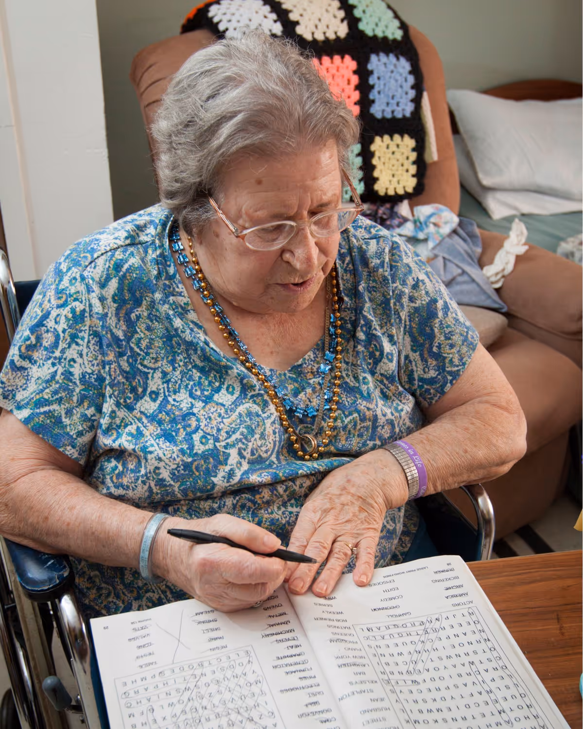 An elderly woman wearing glasses and a blue patterned shirt is sitting in a wheelchair at a table, holding a pen and working on a word search puzzle in a book. She is wearing multiple necklaces and wristbands. Behind her, there is a bed with a colorful crocheted blanket and pillows.