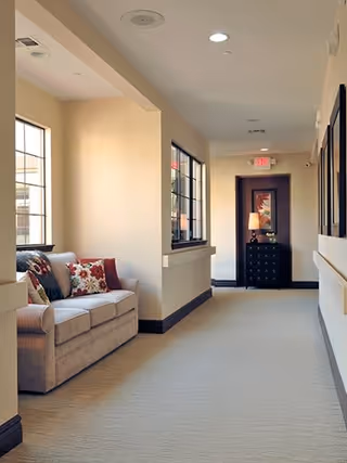 A well-lit hallway with beige walls and carpet, featuring a beige sofa with colorful cushions near large windows on the left side. At the end of the hallway, there is a small black chest of drawers with a lamp and framed artwork above it.