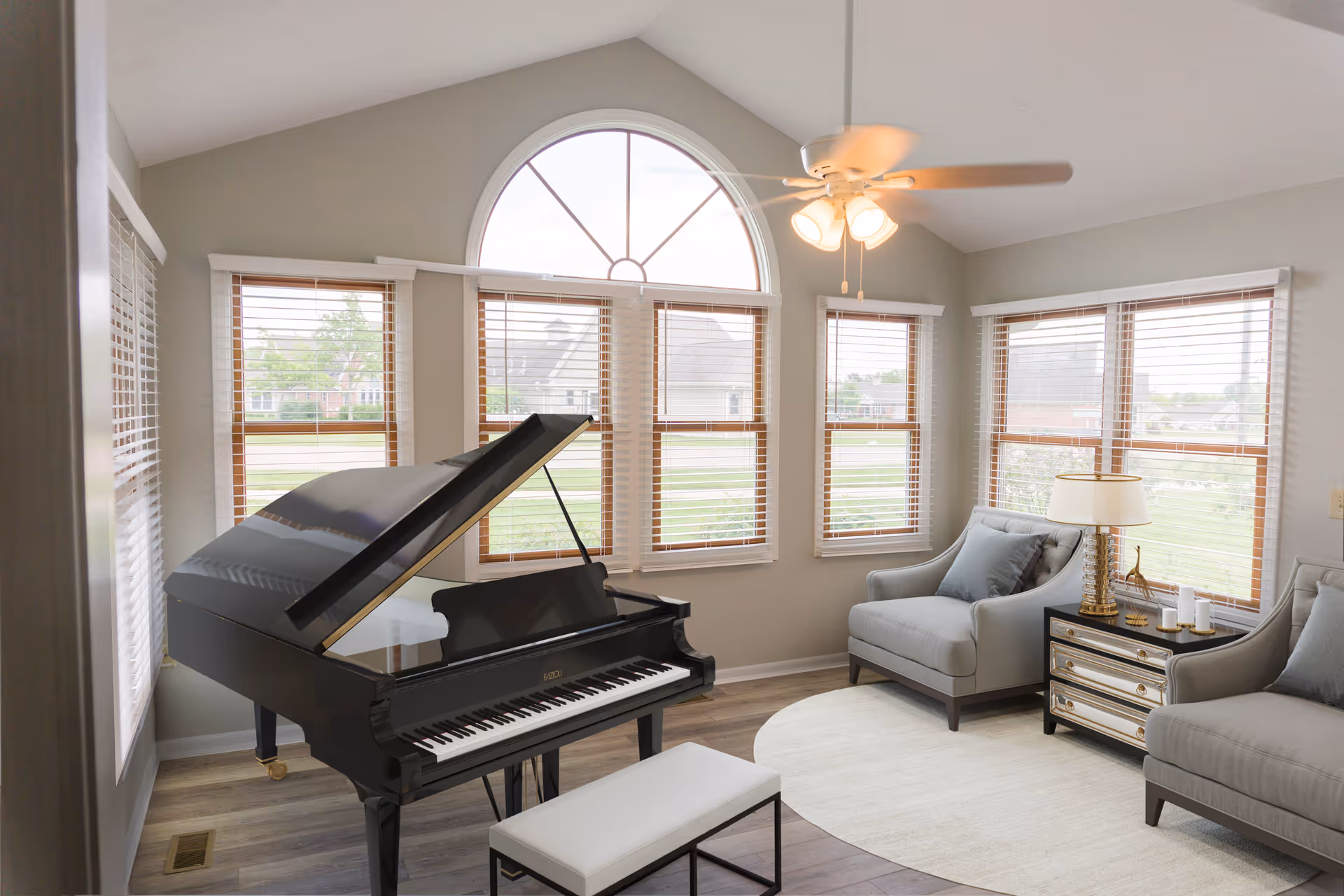 A bright living room with a black grand piano and matching bench in the foreground. The room features large windows with wooden blinds, a ceiling fan with lights, two gray armchairs with blue cushions, a mirrored side table with a lamp and decorative items, and a light-colored circular rug on wooden flooring.