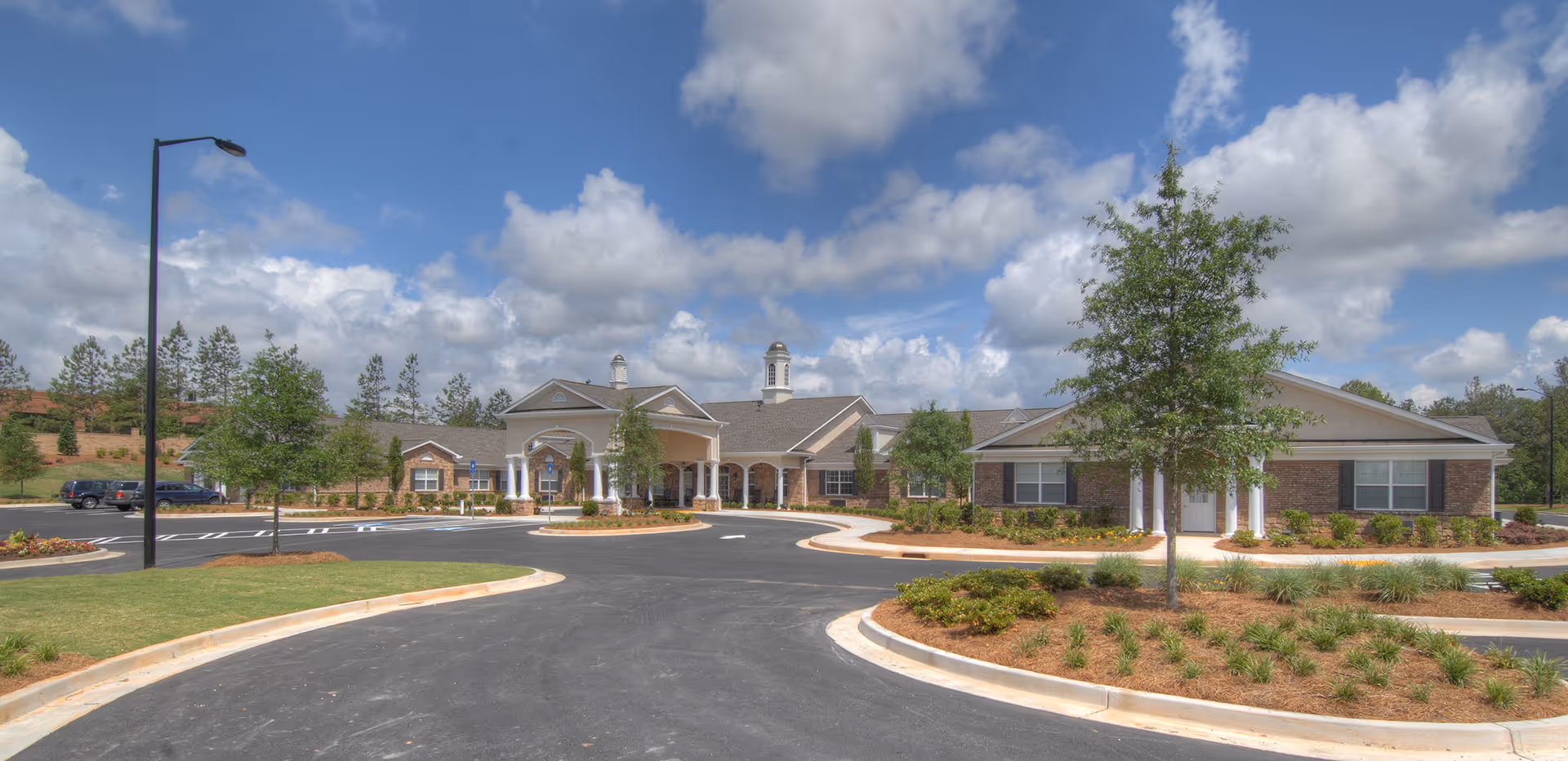 Front exterior view of Benton House of Douglasville, a single-story brick building with white columns and a covered entrance, surrounded by landscaped greenery and a parking lot under a partly cloudy blue sky.