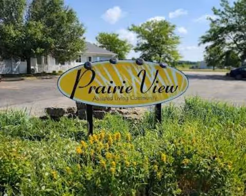 Entrance sign reading 'Prairie View Assisted Living Community' surrounded by plants with the facility building and parking lot visible behind.