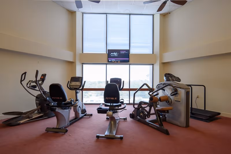 A fitness room with various exercise equipment including stationary bikes, an elliptical machine, a rowing machine, and a treadmill. The room has large windows letting in natural light and a wall-mounted TV above the windows.