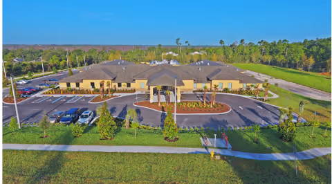 Aerial view of a single-story senior living facility named Gold Choice ALF surrounded by parking lots, landscaped greenery, and trees with a clear sky in the background.