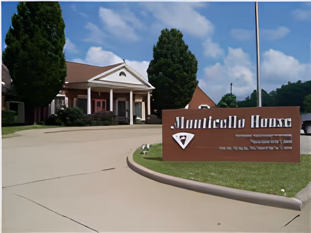 Exterior view of Monticello House facility with a driveway leading to a building with white columns and a triangular pediment. There are tall green trees and a clear blue sky with some clouds. A brown sign with white lettering reads 'Monticello House' near the driveway.