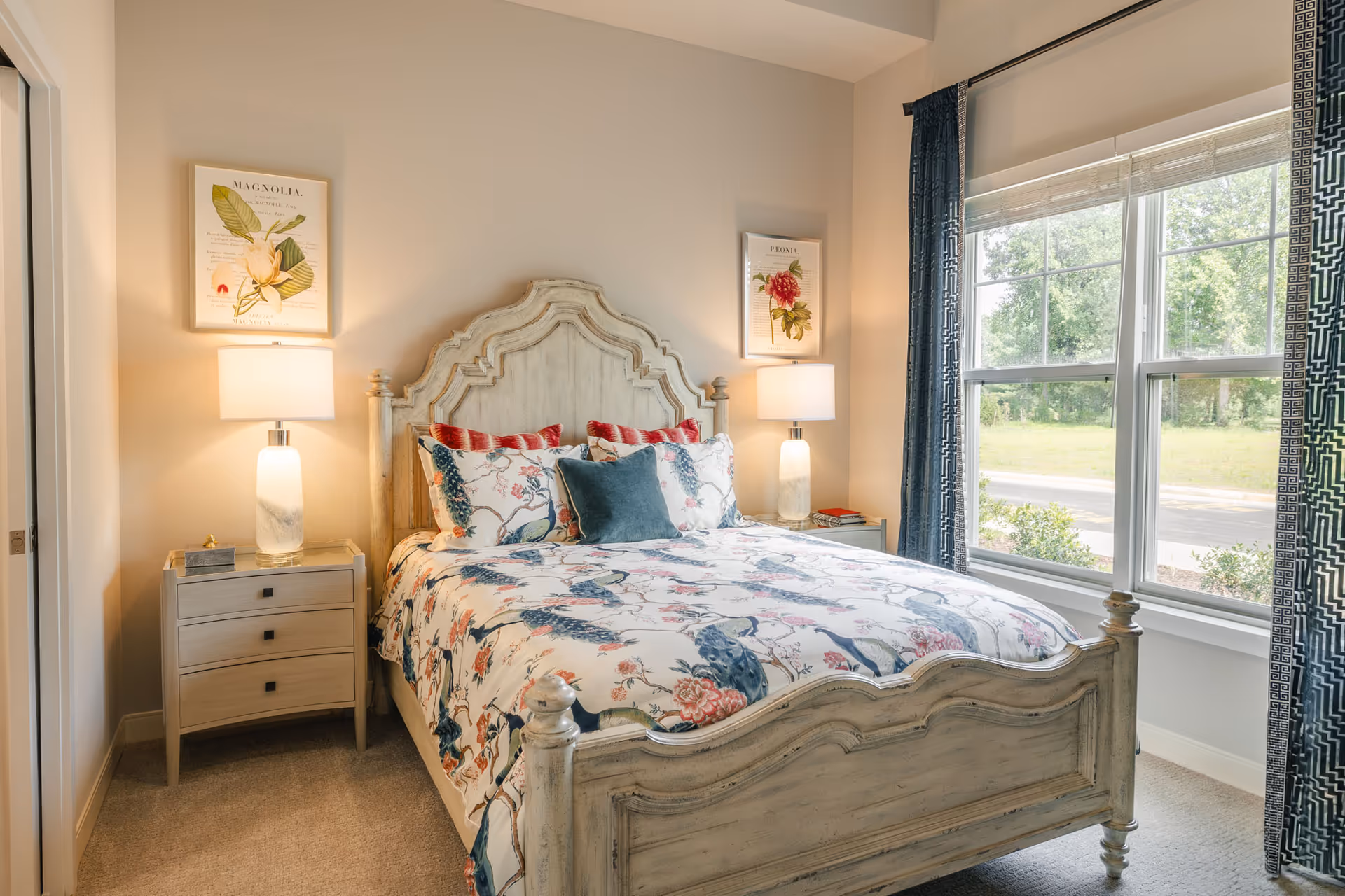 A cozy bedroom with a vintage-style wooden bed featuring a floral and bird patterned bedspread and multiple pillows. There are two bedside tables, each with a modern lamp and framed botanical prints hanging above them. A large window with blue patterned curtains lets in natural light and offers a view of greenery outside.