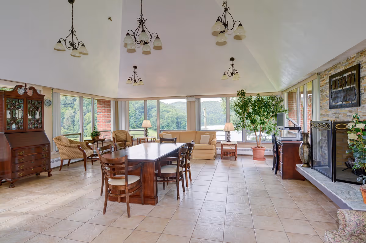 A spacious and bright living room with large windows offering a scenic view of trees and hills. The room features a long wooden dining table with chairs, a beige sofa, armchairs, a wooden cabinet, potted plants, a fireplace with a decorative screen, and multiple hanging light fixtures on a high vaulted ceiling.