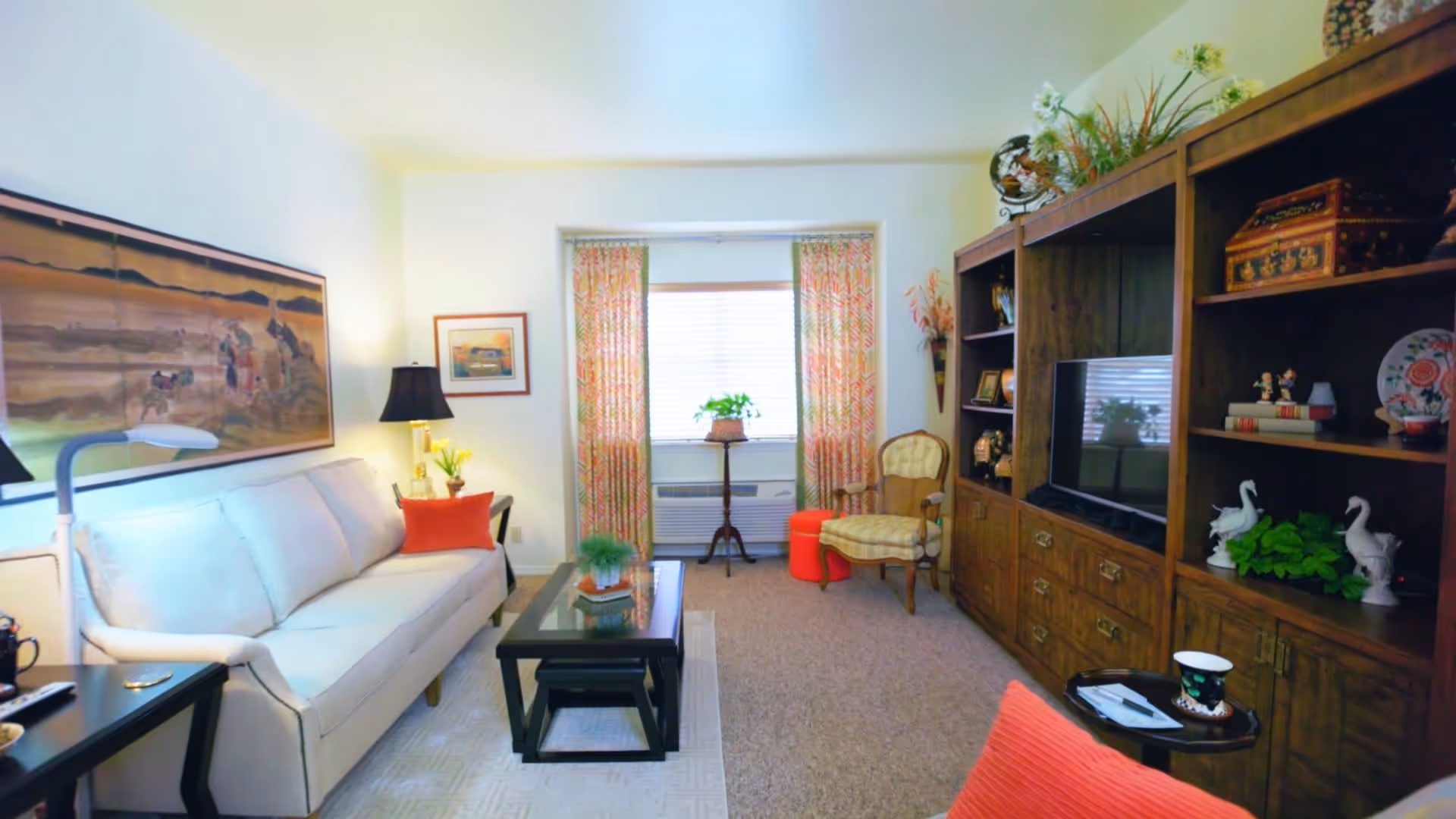 A cozy living room with a white sofa adorned with orange pillows, a black coffee table with a small plant, a wooden entertainment center with a TV and decorative items, an armchair with a red ottoman, and a window with patterned curtains letting in natural light.