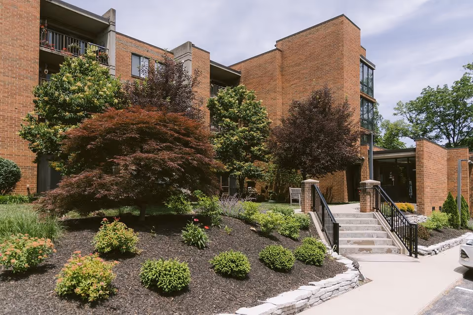 Exterior view of a brick building with landscaped garden featuring various shrubs and trees. There is a concrete walkway with stairs and black railings leading to the entrance of the building. A parked car is partially visible on the right side.