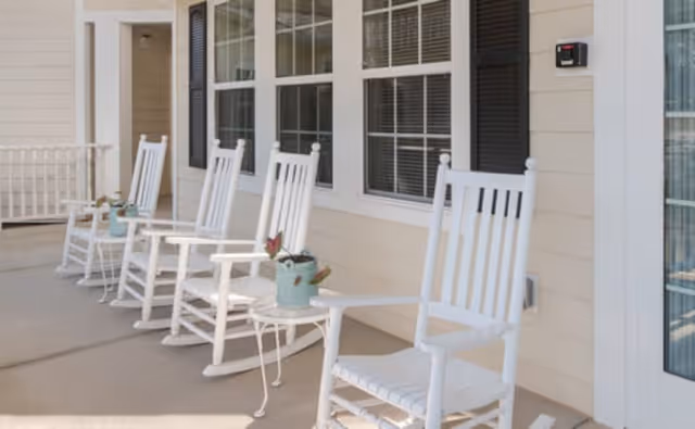 A row of white wooden rocking chairs on a covered porch outside a building with beige siding and black window shutters. Small tables with potted plants are placed between the chairs.