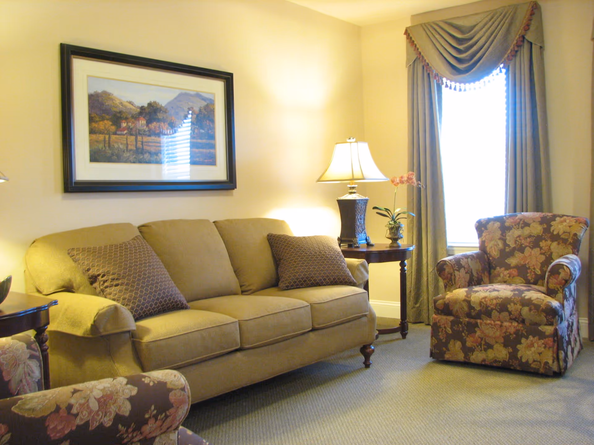 Cozy living room with a beige sofa and pillows, a floral armchair, side table with a lamp, framed artwork, and a curtained window.