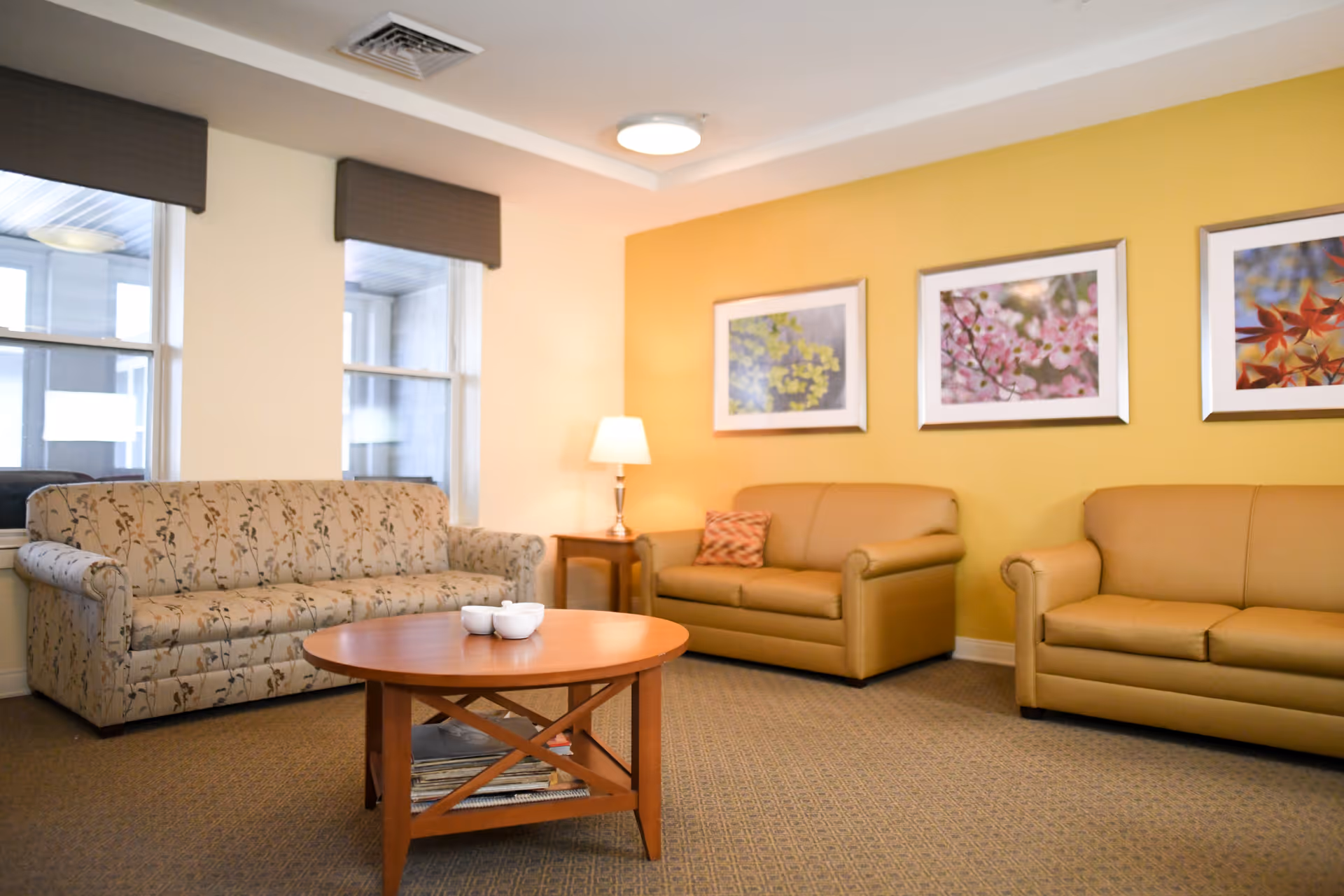 A cozy living room with a floral patterned sofa on the left and two tan leather sofas on the right. A wooden coffee table with three white bowls is in the center. The walls are painted yellow and decorated with three framed pictures of flowers. A table lamp is on a small wooden side table in the corner near the sofas. Two windows with brown valances are on the left wall.