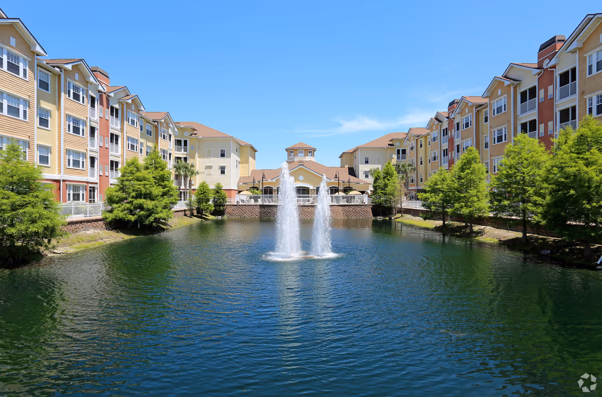 View of a senior living facility named Villa Grande on Saxon with two large multi-story buildings on either side of a pond featuring two water fountains. The buildings have yellow and red exterior walls with multiple windows and balconies. Trees line the edge of the pond under a clear blue sky.