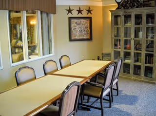 A dining room with a long rectangular table surrounded by eight chairs. On the wall above the table are three decorative stars and a framed Route 66 map. To the right is a glass-front cabinet filled with various items. A window on the left side reflects part of the room.