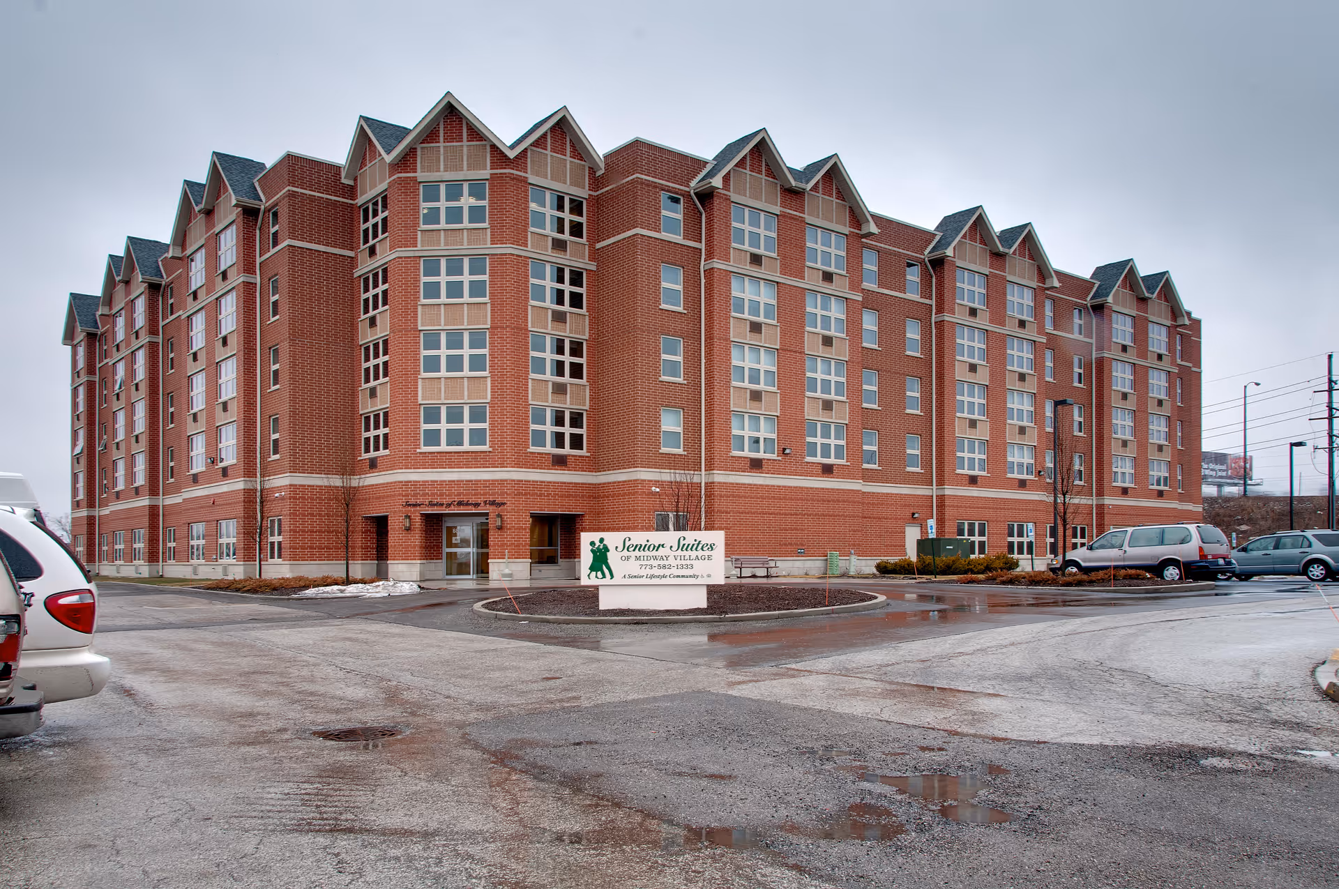 Exterior view of a large multi-story brick building with many windows, identified as Senior Suites of Midway Village. The building is surrounded by a parking lot with several parked cars and a sign in front displaying the facility's name and contact information. The sky is overcast and the ground appears wet.