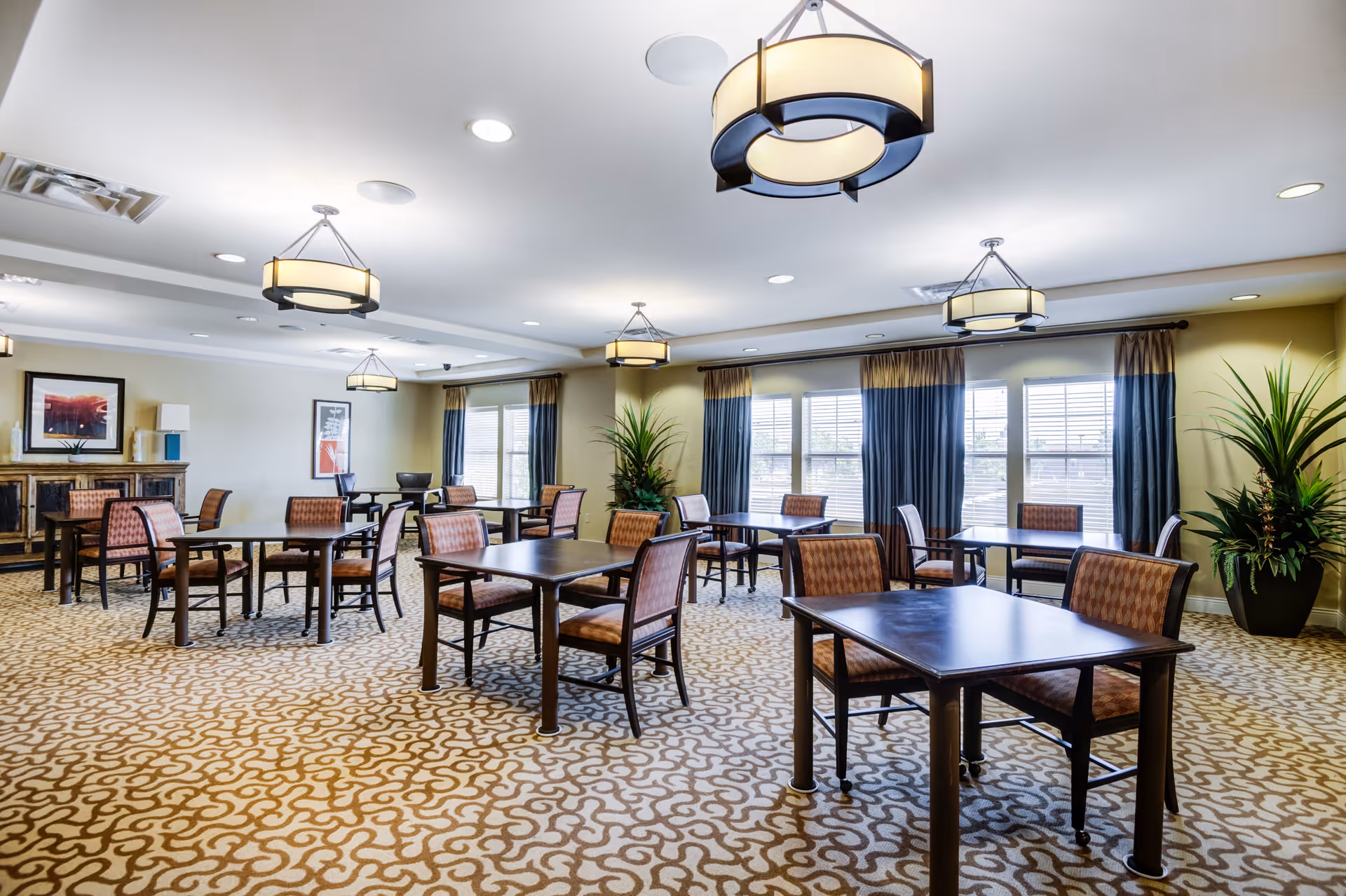A spacious dining room with multiple dark wood tables and chairs with patterned upholstery. The room has large windows with blue and beige curtains, patterned carpet, modern ceiling lights, and decorative plants in the corners. Artwork and a sideboard are visible against the far wall.